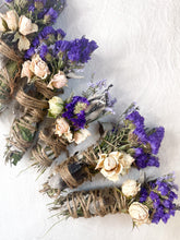 Load image into Gallery viewer, May include: A set of four smudge sticks wrapped in twine and decorated with dried flowers. Each stick features a different crystal, including clear quartz, amethyst, and smoky quartz. The flowers are a mix of purple and white.