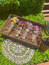 Load image into Gallery viewer, May include: Four bundles of dried herbs and flowers tied with twine and adorned with crystals, presented in a brown box with shredded paper. The bundles are arranged in a row and are surrounded by green foliage.