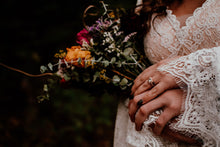 Load image into Gallery viewer, May include: A bride holds a bouquet of flowers with orange, pink, and purple blooms. The bouquet is wrapped in greenery and has a rustic, natural feel. The bride is wearing a white lace dress and has a diamond ring on her finger.