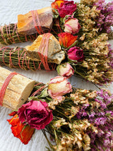 Load image into Gallery viewer, May include: Three bundles of dried herbs and flowers tied with coral-colored string. Each bundle features a unique stone or wood piece secured to the top, accompanied by dried roses in shades of red, pink, and orange, and sprigs of purple statice.
