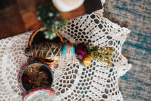 Load image into Gallery viewer, May include: A bundle of dried herbs and flowers wrapped in twine with a turquoise stone, positioned next to a round, colorful metal tin containing a candle with embedded botanical elements, all resting on a white crocheted lace doily.