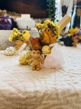 Load image into Gallery viewer, May include: A small, smiling troll figurine with a light brown body and yellow hair. The troll is wearing a flower crown made of dried roses and other flowers. The crown is sitting on a large, pink, rose quartz crystal.