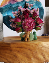 Load image into Gallery viewer, May include: A small, brown troll doll figurine with a pink flower crown made of dried flowers. The doll is sitting on a wooden surface with green stones around it.