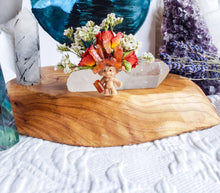 Load image into Gallery viewer, May include: A small orange troll figurine with a crown of carnelian stones and white flowers on a wooden base with clear quartz crystals.