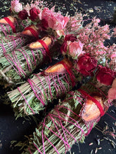 Load image into Gallery viewer, May include: Close-up of several bundles of dried herbs, sage, and flowers. Each bundle is wrapped with pink string and red ribbon. The bundles are adorned with dried roses and other pink and red flowers. The background is black.