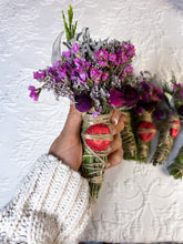 Load image into Gallery viewer, May include: A bundle of dried flowers and herbs, featuring vibrant purple blooms and green foliage, wrapped with twine and a slice of dried strawberry. Several similar bundles are visible in the background.