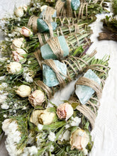 Load image into Gallery viewer, May include: A bundle of dried herbs and flowers tied with twine and adorned with green crystals. The bundle is surrounded by dried roses and other flowers.