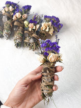 Load image into Gallery viewer, May include: A bundle of dried herbs and flowers wrapped in twine with a clear quartz crystal. The bundle is tied with twine and decorated with dried flowers, including purple statice and white roses.
