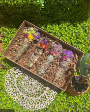 Load image into Gallery viewer, May include: A set of four smudge sticks with dried flowers and crystals. The smudge sticks are wrapped in twine and have a variety of colors, including purple, yellow, and green. The smudge sticks are in a brown box with shredded paper.
