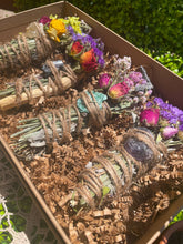 Load image into Gallery viewer, May include: A set of four smudge sticks with various crystals and dried flowers. The smudge sticks are wrapped in twine and are in a brown box with shredded paper.
