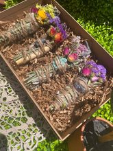 Load image into Gallery viewer, May include: A set of four smudge sticks in a brown box with shredded paper. Each smudge stick is wrapped in twine and includes dried flowers and crystals. The crystals are amethyst, clear quartz, green aventurine, and black tourmaline.
