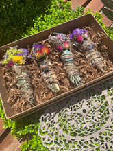 Load image into Gallery viewer, May include: Four smudge sticks with dried flowers and crystals in a brown box. The smudge sticks are wrapped in twine and have different colored crystals at the end. The box is filled with brown packing paper.
