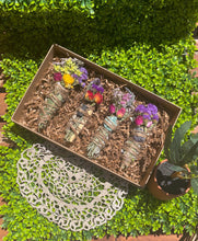 Load image into Gallery viewer, May include: A set of four smudge sticks with dried flowers and crystals in a brown box. The smudge sticks are wrapped in twine and have different colored flowers and crystals.
