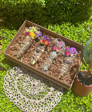 Load image into Gallery viewer, May include: Four bundles of dried herbs and flowers tied with twine, each containing a crystal. The bundles are arranged in a brown box lined with brown paper shreds. The box is sitting on a white crocheted doily.
