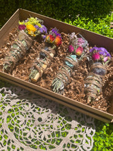 Load image into Gallery viewer, May include: Four smudge sticks with crystals and dried flowers in a brown box. The smudge sticks are wrapped in twine and have different colored crystals and flowers.
