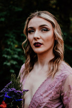 Load image into Gallery viewer, May include: A woman with long blonde hair wearing a pink lace top with a plunging neckline. She is looking at the camera with a serious expression. She is holding a bouquet of purple flowers in her left hand.
