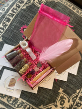 Load image into Gallery viewer, May include: A pink and brown gift box filled with crystals, herbs, a feather, a candle, and a small pouch. The box is surrounded by small cards with words like "abundance", "strength", "protection", "confidence", and "rose quartz".

