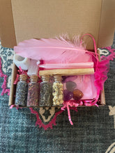 Load image into Gallery viewer, May include: A pink gift box filled with various items, including a pink feather, a white candle, four small glass jars with dried herbs and flowers, and several purple and orange gemstones. The box is lined with pink tissue paper and has a pink ribbon.
