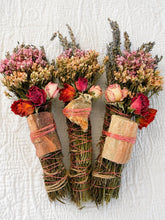 Load image into Gallery viewer, May include: Three bundles of dried herbs and flowers, each featuring pink and orange dried roses, lavender sprigs, and statice. Each bundle is wrapped with twine and secured with a piece of wood or a stone, tied with thin pink string.
