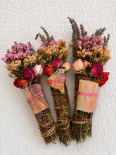 Load image into Gallery viewer, May include: Three bundles of dried herbs and flowers, including lavender, pink statice, and red roses. Each bundle is wrapped with twine and features a unique, flat, polished stone secured to the front with thin pink string.
