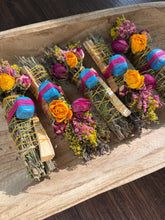 Load image into Gallery viewer, May include: Four bundles of dried herbs and palo santo wood sticks wrapped with yellow twine. Each bundle is decorated with dried yellow roses, pink rosebuds, small purple flowers, and a single bright blue stone secured with pink thread.
