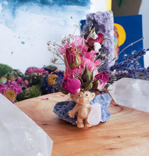 Load image into Gallery viewer, May include: A small, brown troll figurine with a pink flower crown made of dried flowers. The troll is holding a pink crystal in its hand. The figurine is sitting on a blue rock.
