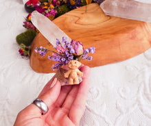 Load image into Gallery viewer, May include: A small, light brown troll doll figurine with a purple flower crown sits on a clear quartz crystal. The crystal is surrounded by dried purple flowers.
