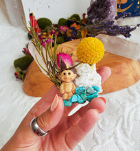 Load image into Gallery viewer, May include: A small, smiling troll figurine surrounded by turquoise stones, a white crystal, a dried yellow flower, and a pink rose bud. The figurine is sitting on a bed of turquoise stones.
