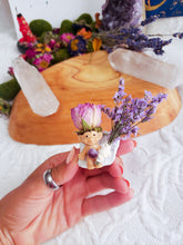 Load image into Gallery viewer, May include: A small, smiling troll figurine with a purple flower crown sits on a white rock with a bouquet of lavender flowers. The troll is holding a purple gemstone.
