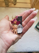 Load image into Gallery viewer, May include: A small plastic troll figurine with a clear quartz crystal, a dried rose, and a black twig in a hand.
