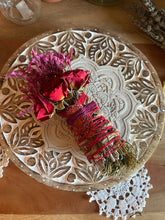 Load image into Gallery viewer, May include: A handcrafted smudge stick with dried red roses and purple flowers, wrapped with red and purple twine. The bundle rests on a carved wooden plate with a white mandala design, and a white lace doily.
