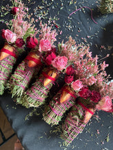 Load image into Gallery viewer, May include: Several bundles of dried herbs and flowers are arranged on a black surface. Each bundle is wrapped with pink string and red ribbon, and topped with pink and red roses. The bundles are a mix of green, pink, and red.
