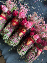 Load image into Gallery viewer, May include: Close-up of several bundles of dried herbs, wrapped with pink string and adorned with dried roses and other flowers. The bundles are green and brown, with red and pink accents. The background is a dark, textured surface.
