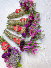 Load image into Gallery viewer, May include: Bundles of dried sage, adorned with dried strawberries and various purple and pink flowers, are tied with twine. The bundles are arranged on a white, textured surface.

