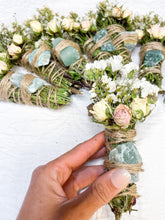 Load image into Gallery viewer, May include: A bundle of dried herbs and flowers tied with twine and adorned with a green crystal. The bundle is held in a hand, and the flowers include white baby's breath and pink roses.
